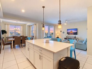 A modern kitchen with a central island, dining area with chairs, and a living room with a sofa and TV.