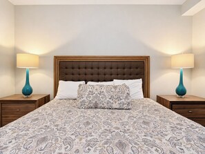 A bedroom with a tufted headboard, two bedside tables with lamps, a patterned bedspread, and a nightstand drawer.