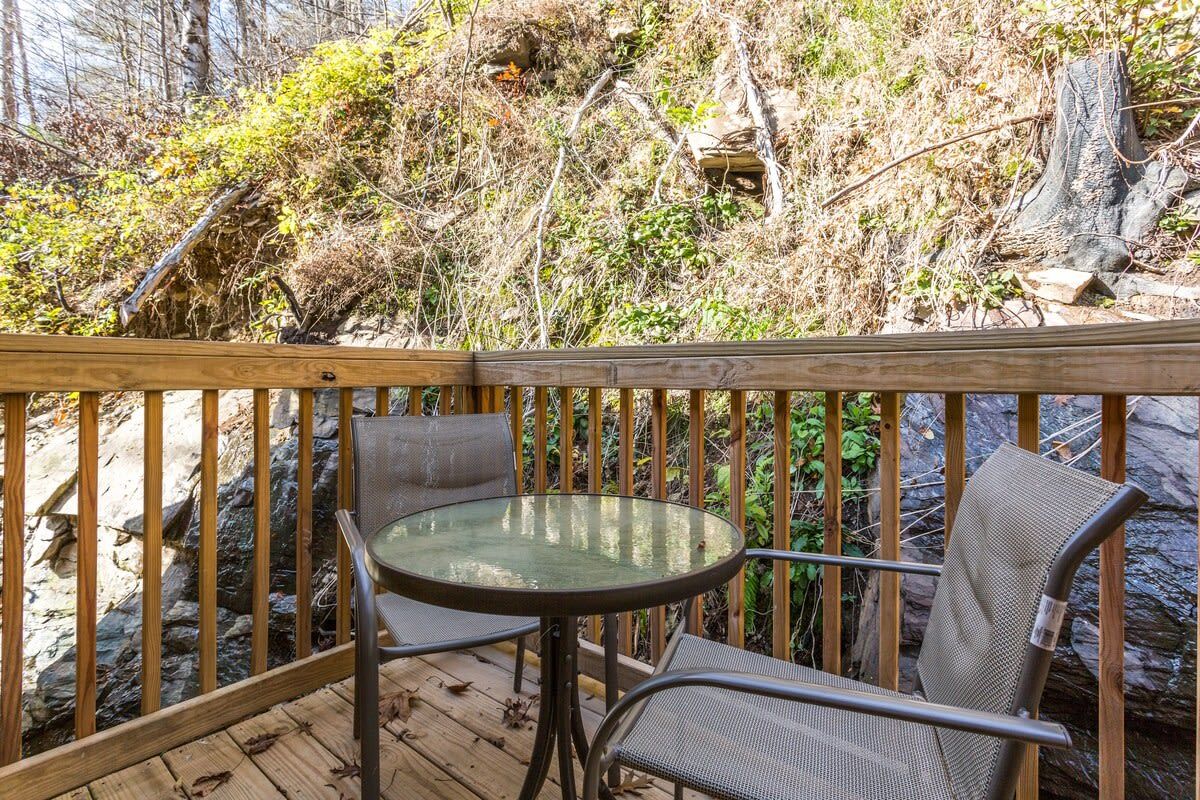 Eine Holzterrasse mit einem Glastisch und Metallstühlen, umgeben von natürlicher Vegetation und einer felsigen Landschaft.