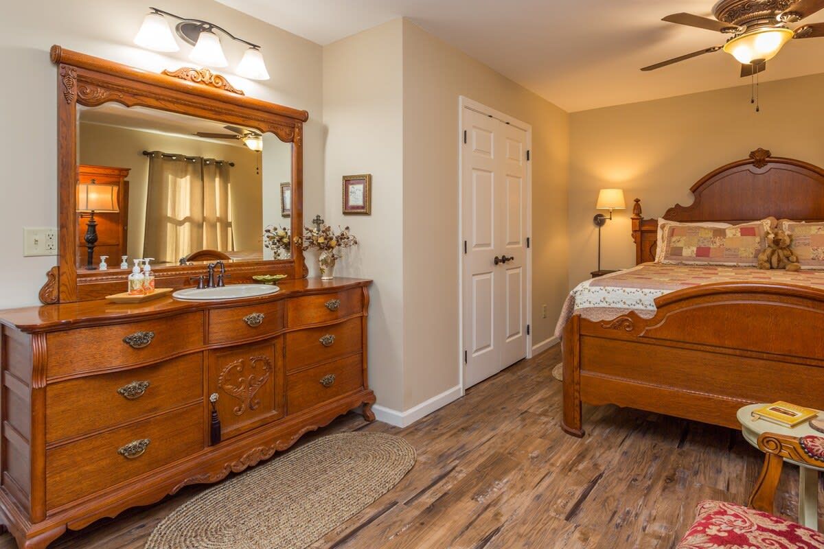 A bedroom with a wooden dresser, a large mirror, a bed with a headboard, a ceiling fan, and a nightstand.