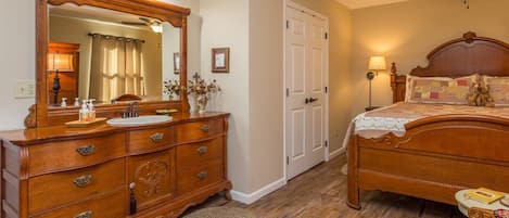 A bedroom with a wooden dresser, a large mirror, a bed with a headboard, a ceiling fan, and a nightstand.