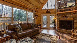 Interior acogedor de una cabaña con chimenea de piedra, mobiliario de madera y ventanas amplias con vistas al bosque circundante.