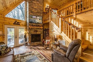Interior acogedor de una cabaña de madera con chimenea, una escalera de madera y un ventanal con vistas a una terraza.