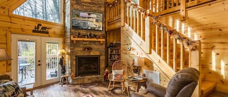 A cozy cabin interior with a fireplace, wooden staircase, and a large window overlooking a deck.