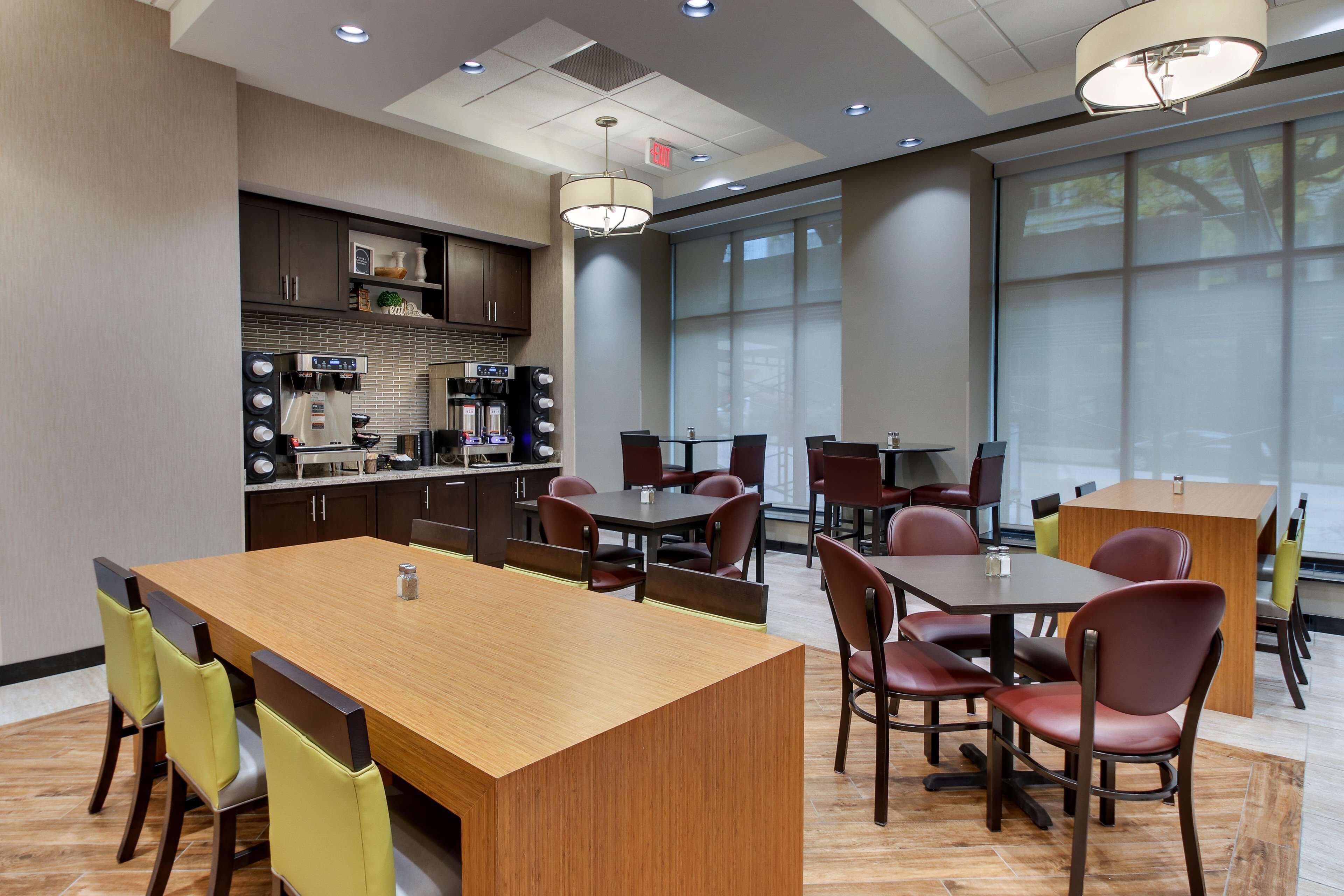 A modern dining area with a central wooden table and surrounding chairs, a coffee station, and large windows providing natural light.