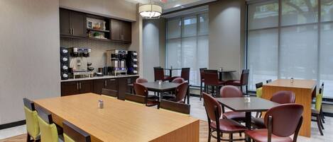 A modern dining area with a central wooden table and surrounding chairs, a coffee station, and large windows providing natural light.