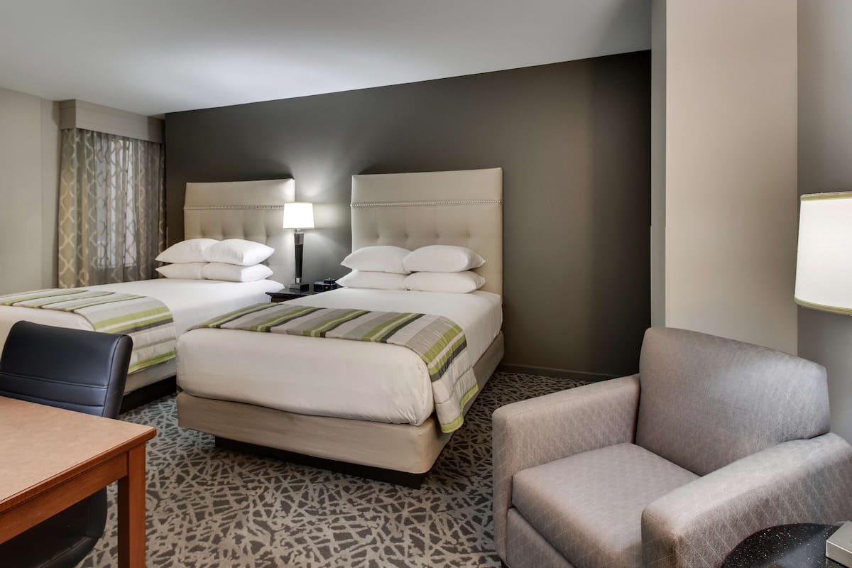 A hotel room with two beds, a desk, a chair, and a lamp.