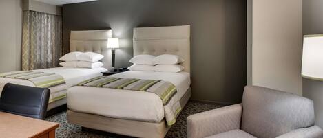 A hotel room with two beds, a desk, a chair, and a lamp.