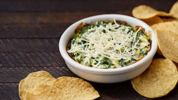 A bowl of spinach dip with shredded cheese, surrounded by tortilla chips on a wooden surface.