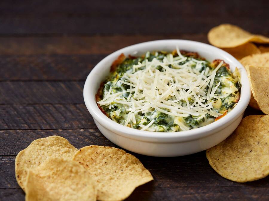 A bowl of spinach dip with shredded cheese, surrounded by tortilla chips on a wooden surface.