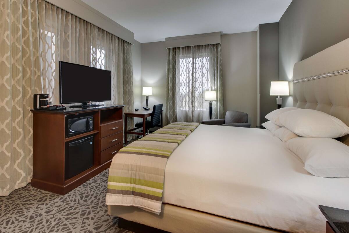 A hotel room with a large bed, a flat-screen TV, a desk with a chair, two lamps, and a minibar.