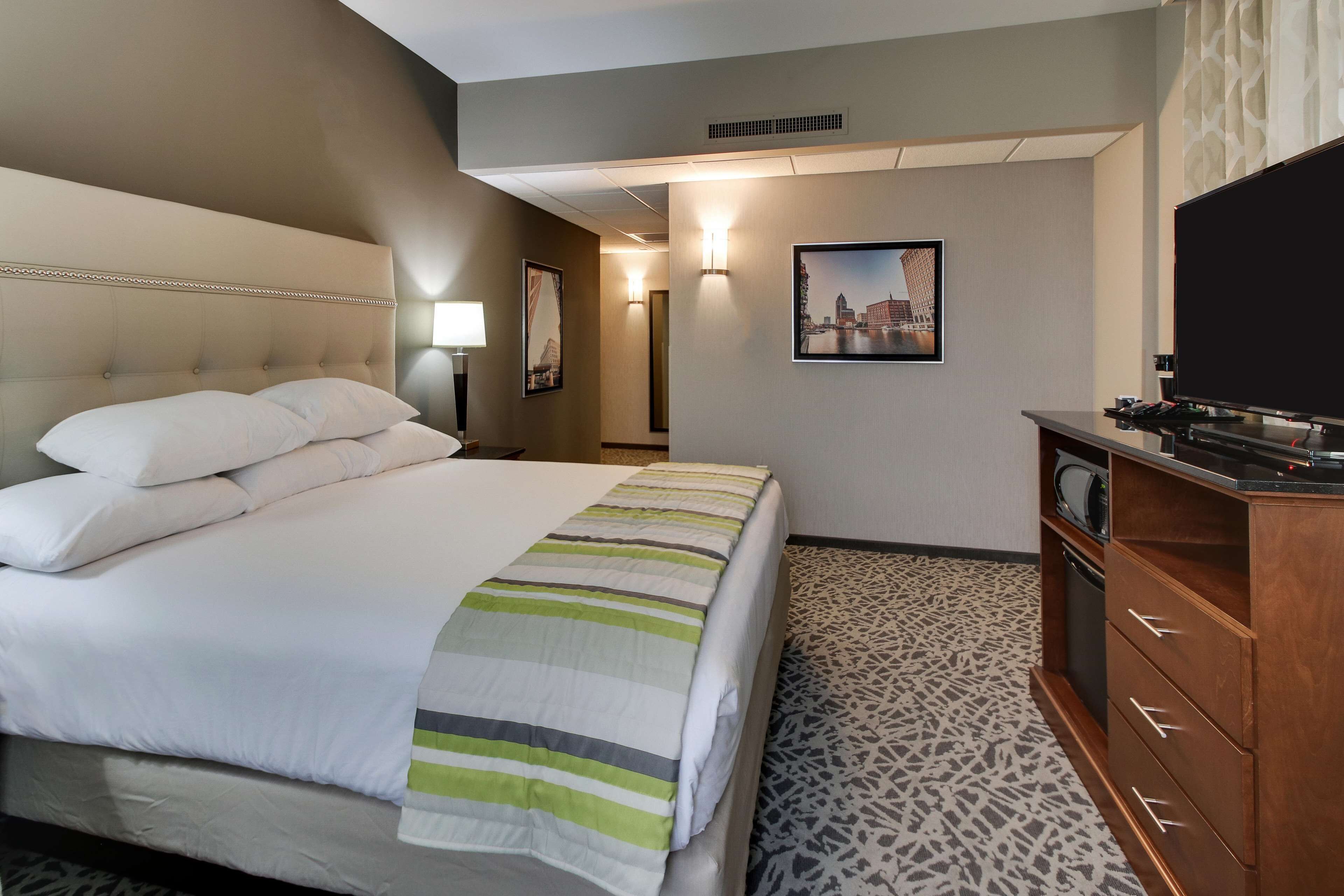 A hotel room with a large bed, a flat-screen TV, a wooden dresser, and a patterned carpet.