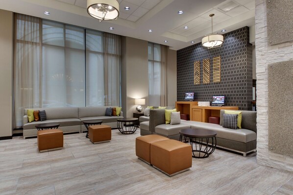 Un moderno vestíbulo de hotel con una zona de estar que incluye sofás, otomanas, una mesa de centro y un televisor montado en una pared con textura.