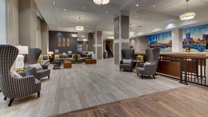 A spacious lobby with a modern design, featuring a seating area with armchairs and sofas, a bar with a counter, and a large cityscape mural on the wall.