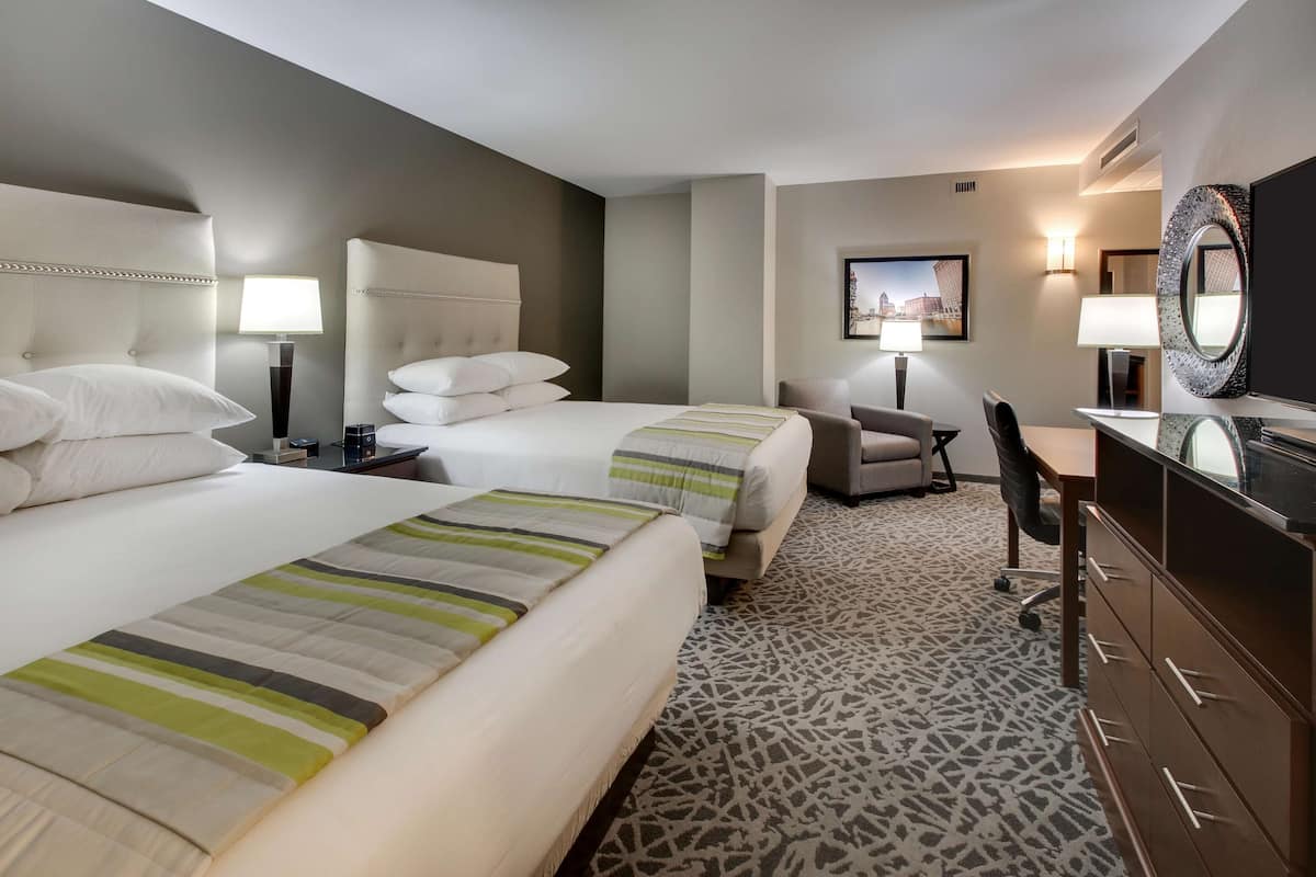 A hotel room with two beds, a desk, a chair, a dresser, and a television.