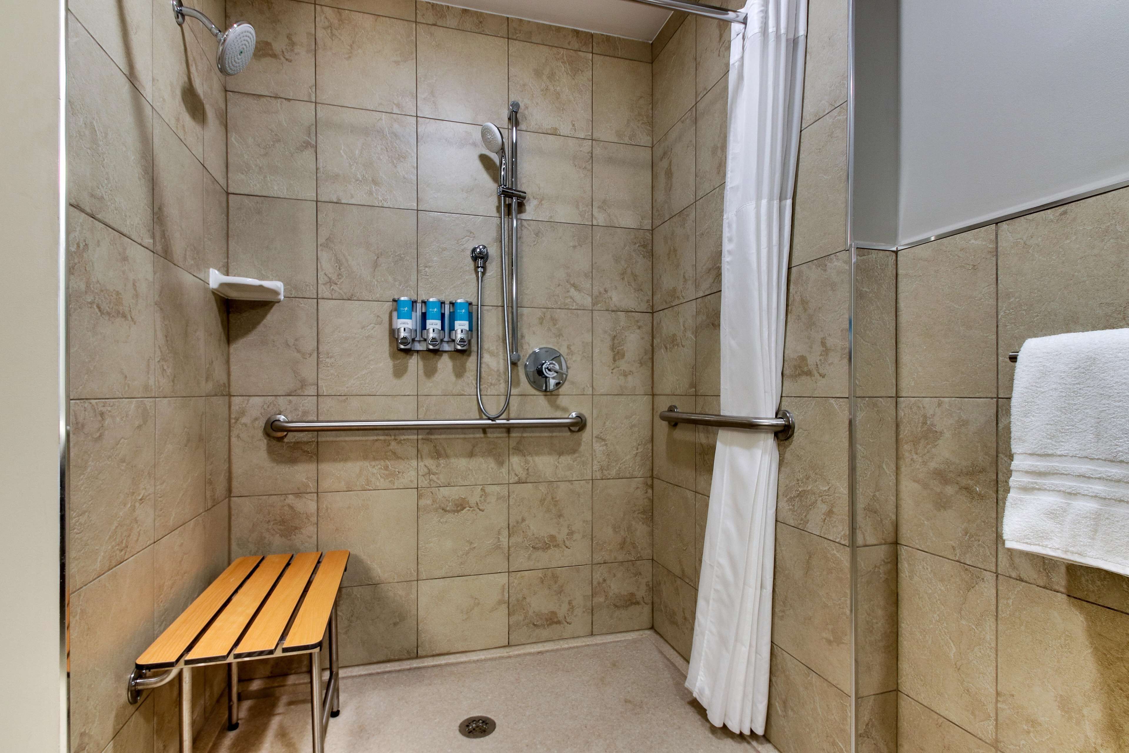 A shower area with beige tiles, a bench, a handrail, a showerhead, a towel rack, and a curtain.