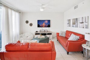 A living room with a red sofa, a blue ottoman, a white bean bag, and a television displaying fireworks.