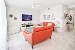 A modern living room with a large orange sofa, a white bean bag, and a flat-screen TV displaying fireworks.