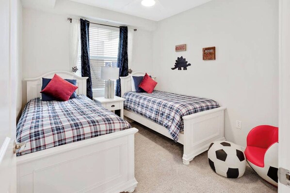 A bedroom with two beds, each with a plaid bedspread and red pillows, a window with blinds, and a red chair with a soccer ball.
