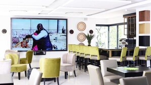 A modern hotel lobby with a large flat-screen TV displaying a news broadcast, comfortable seating, and a dining area with yellow chairs and a wooden table.