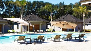 A pool area with lounge chairs and umbrellas, surrounded by buildings with tiled roofs and palm trees.