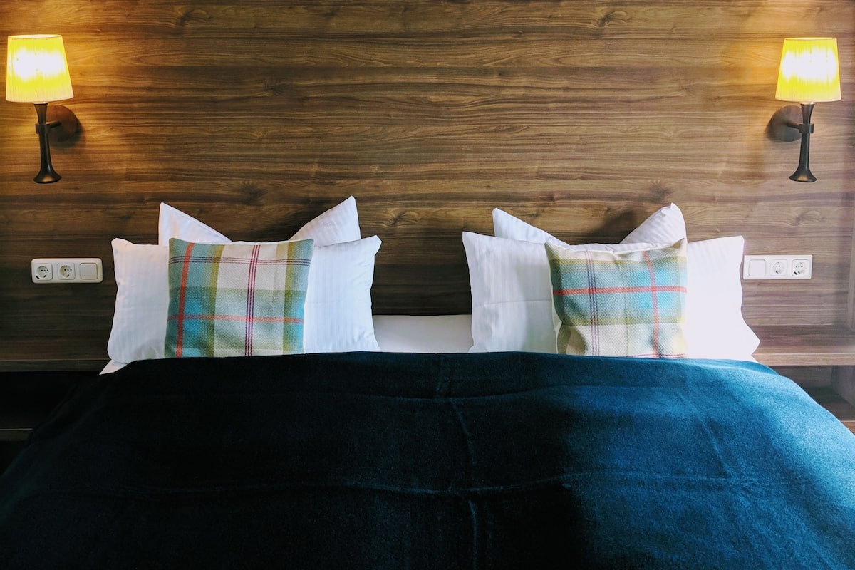 A bed with a dark blue cover, two white pillows, and a plaid pillow. There are two wall-mounted lamps on either side of the bed.