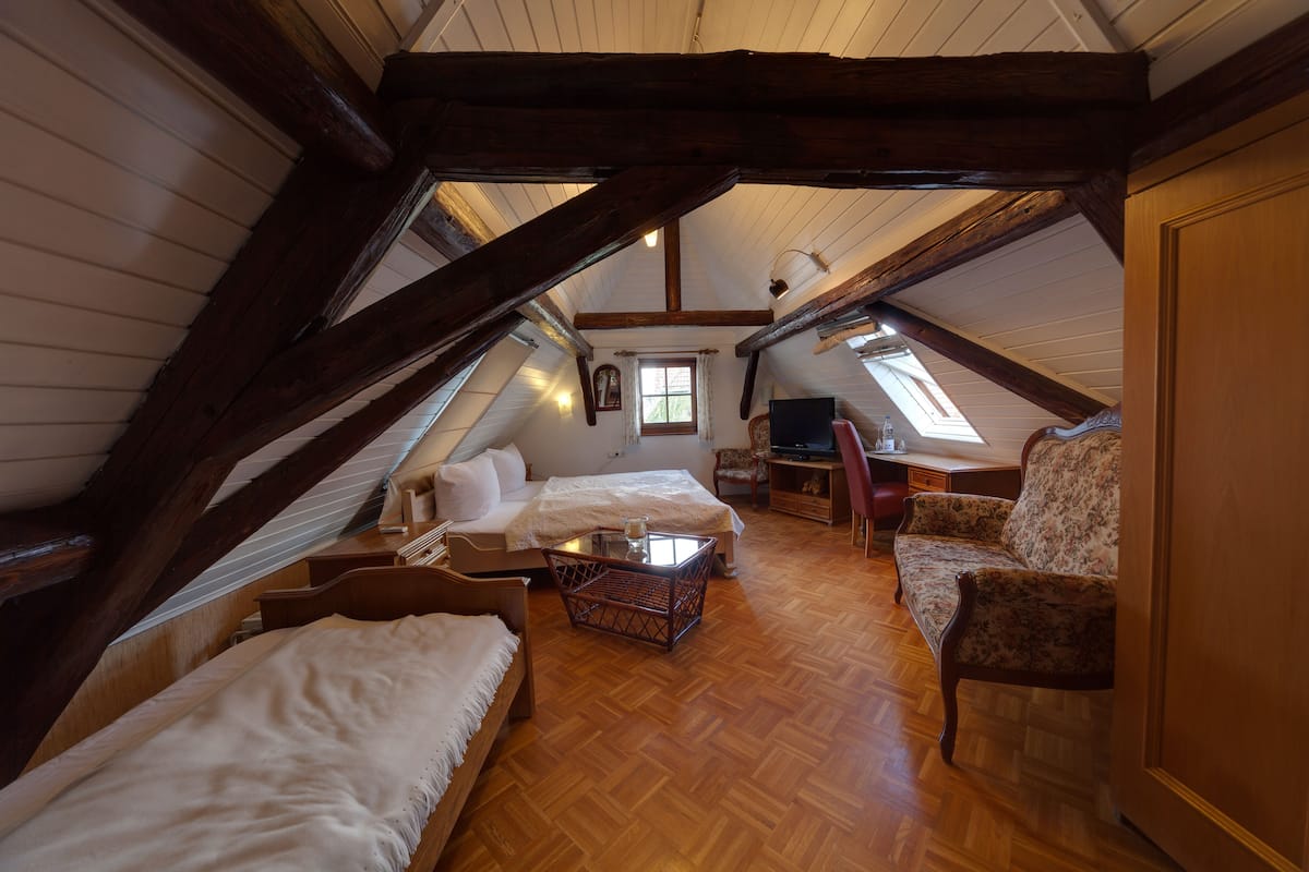 A cozy attic bedroom with wooden beams, a bed, a desk, a chair, and a small window.