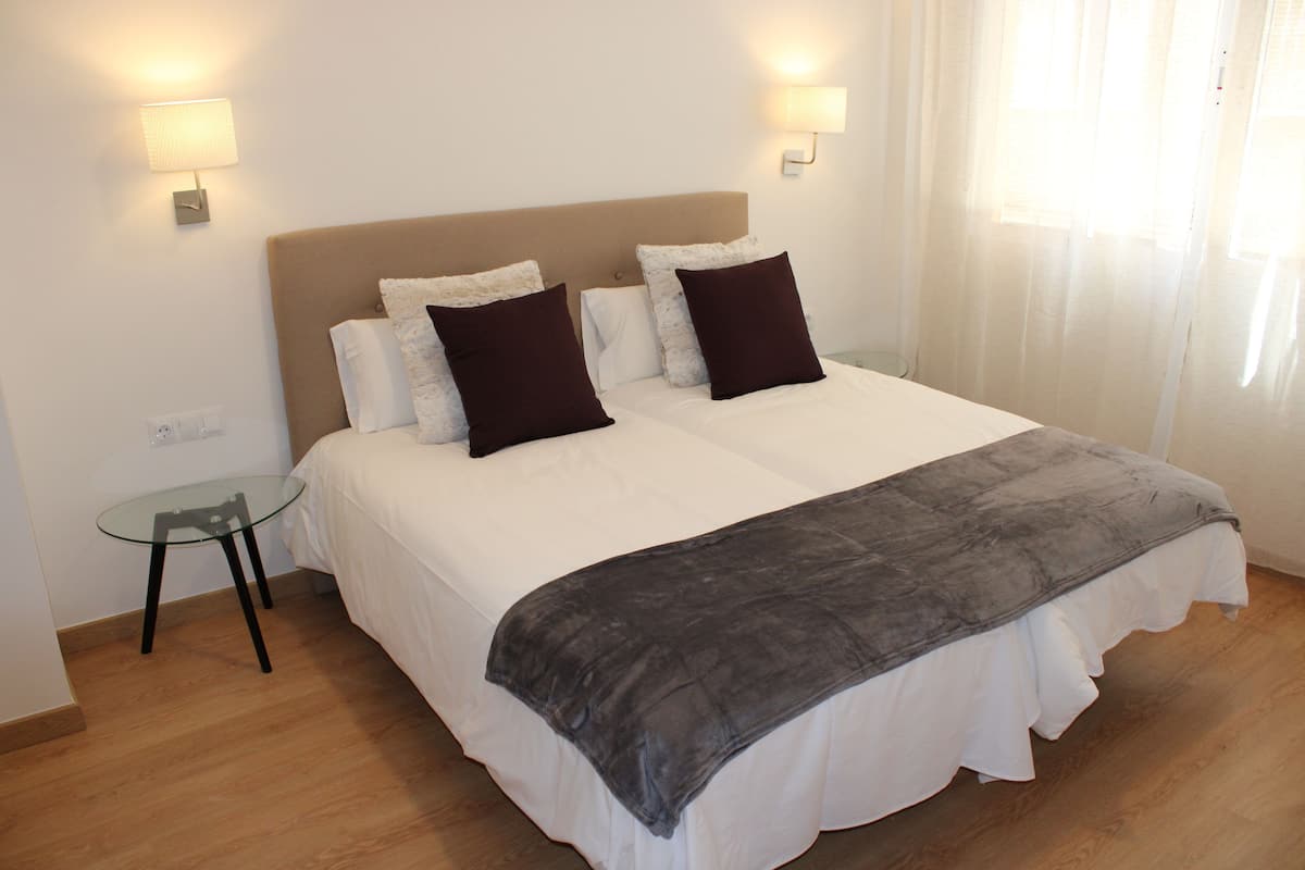 Een net opgemaakt bed met een grijze plaid, twee donkere kussens en een witte dekbedovertrek. Er staat een klein glazen bijzettafeltje naast het bed met een lamp erop.
