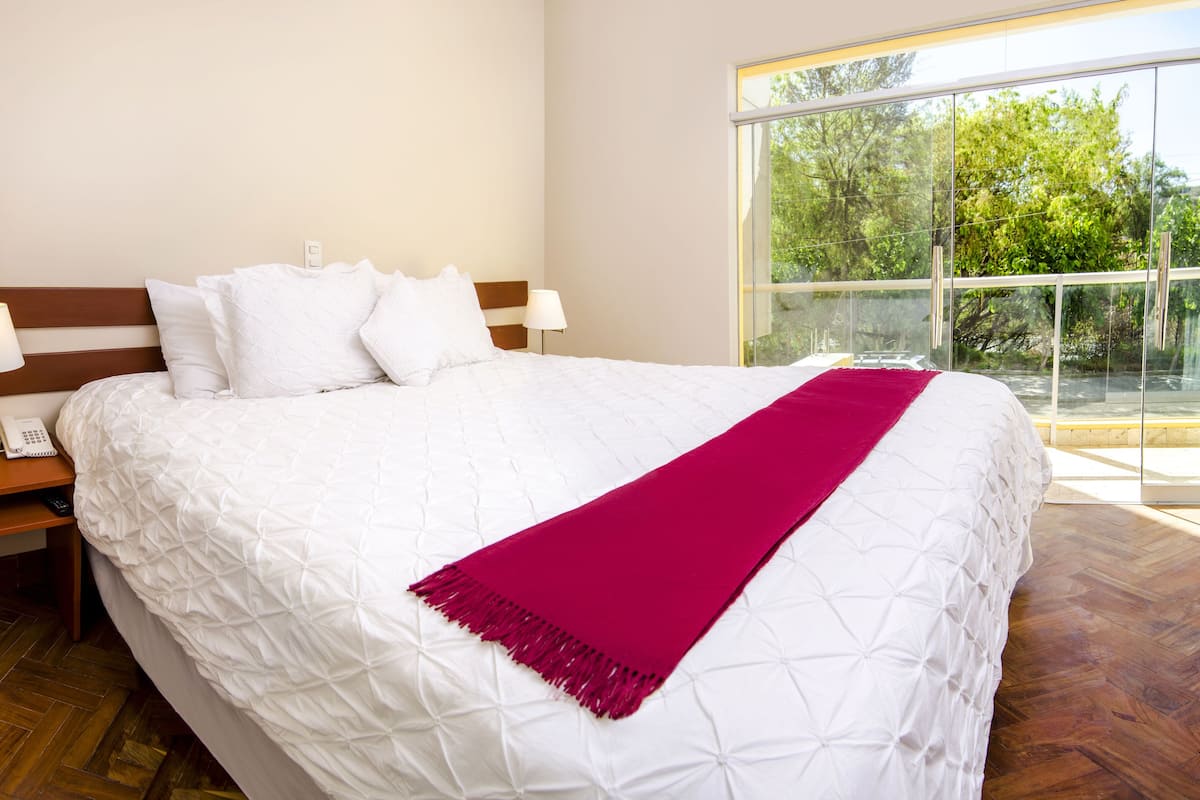 A hotel room with a large bed, a nightstand with a phone and remote, and a window with a view of trees.