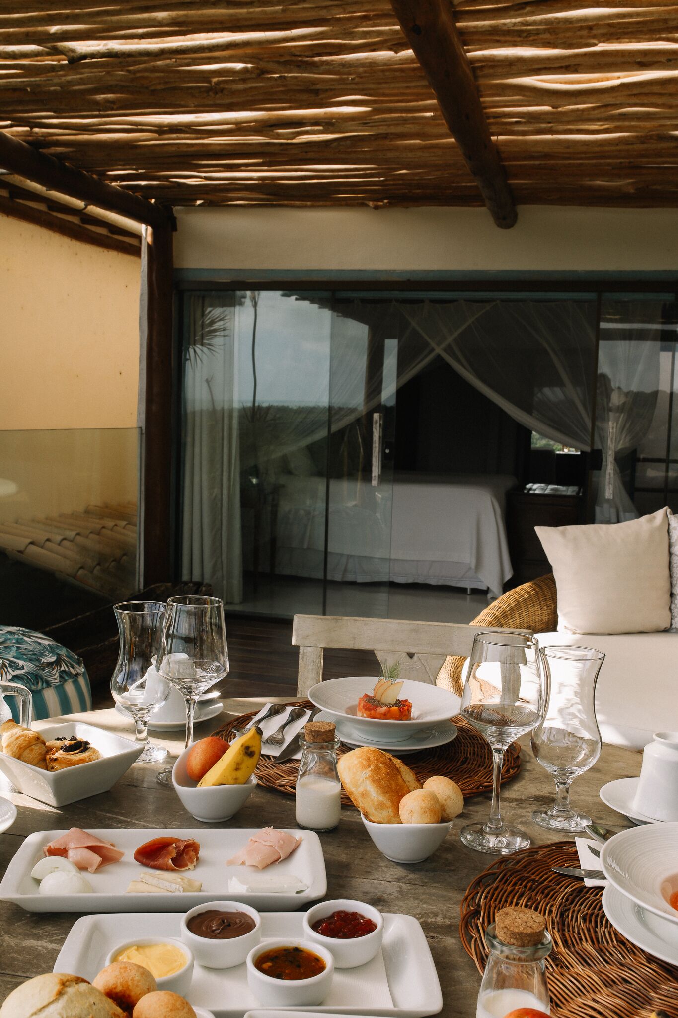 Une table à manger avec divers plats et des verres, une chaise en osier et, au fond, un lit à baldaquin.