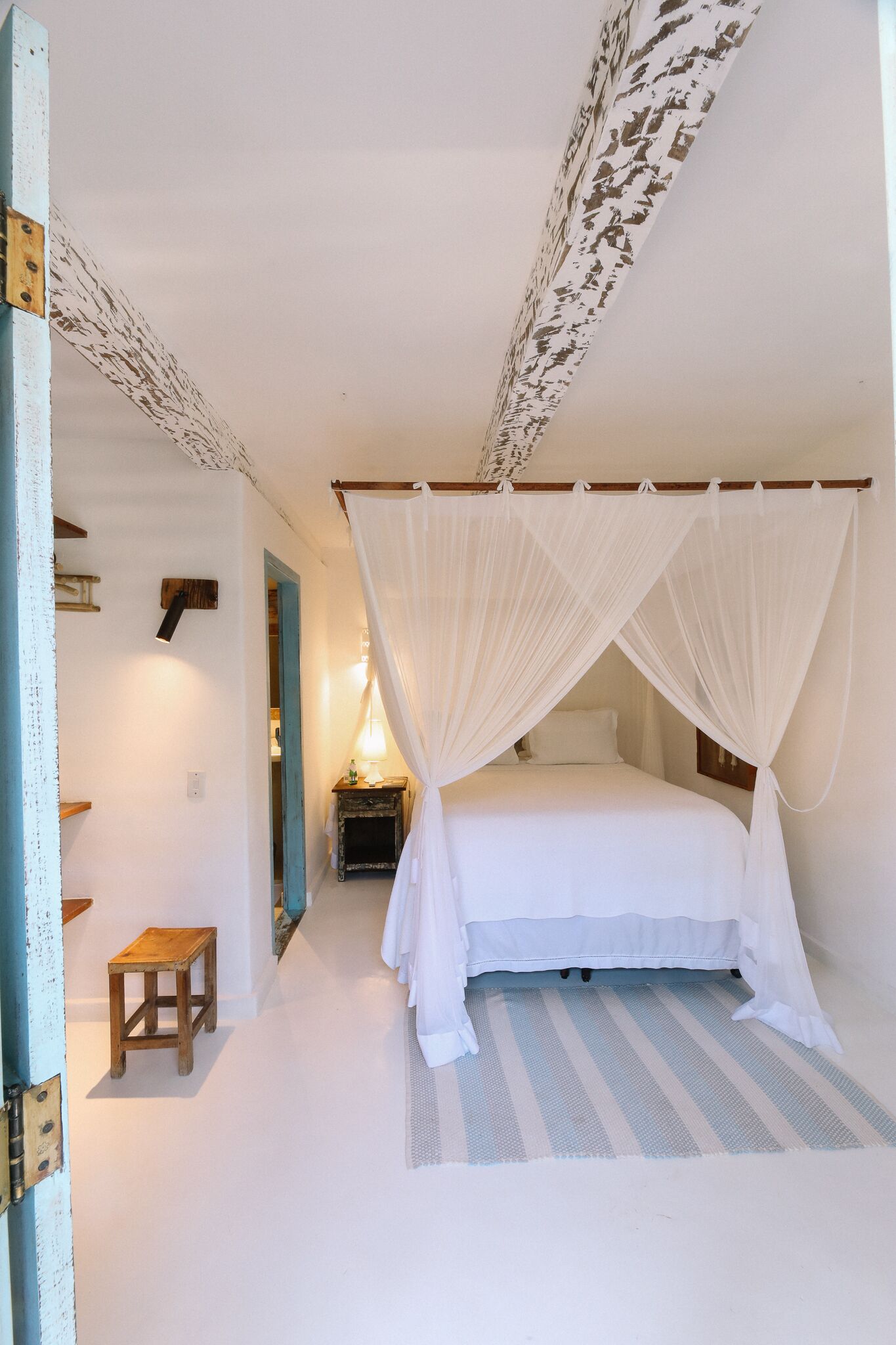 A bedroom with a canopy bed, a small wooden stool, and a nightstand with a lamp.