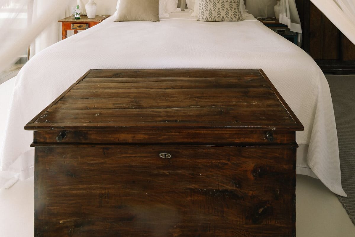 A bedroom with a large bed, white bedding, a wooden chest, and a nightstand with a lamp.