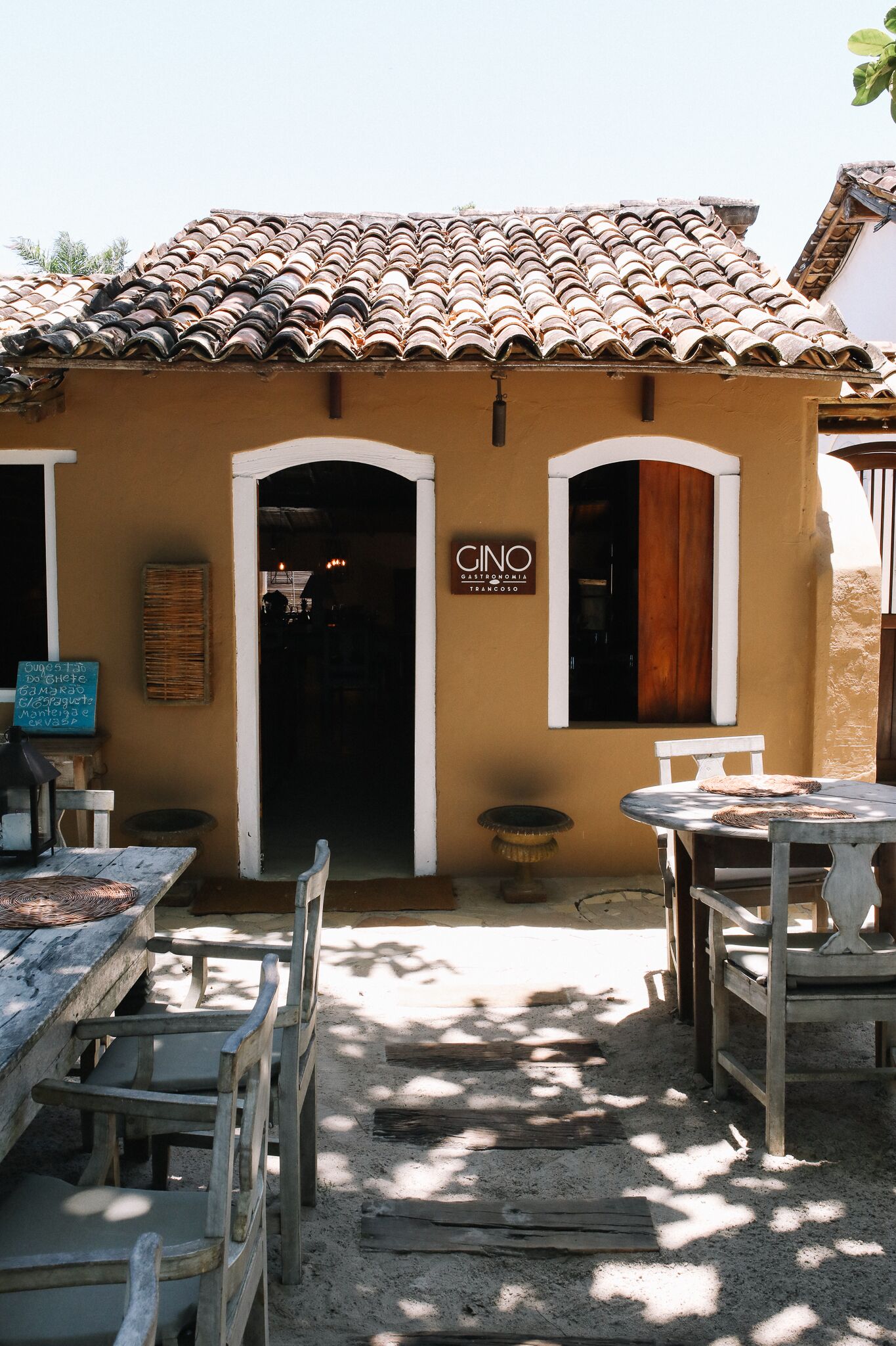 Un bâtiment avec un toit recouvert de tuiles, une enseigne indiquant « Gino », et des tables et des chaises disposées à l’extérieur pour créer un espace de restauration.