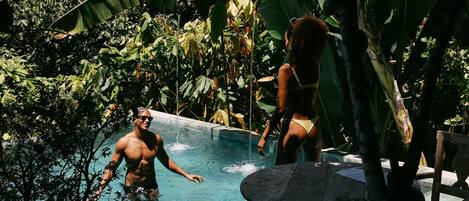 Zona de piscina rodeada de exuberante follaje tropical, con muelle de madera y pared de piedra.