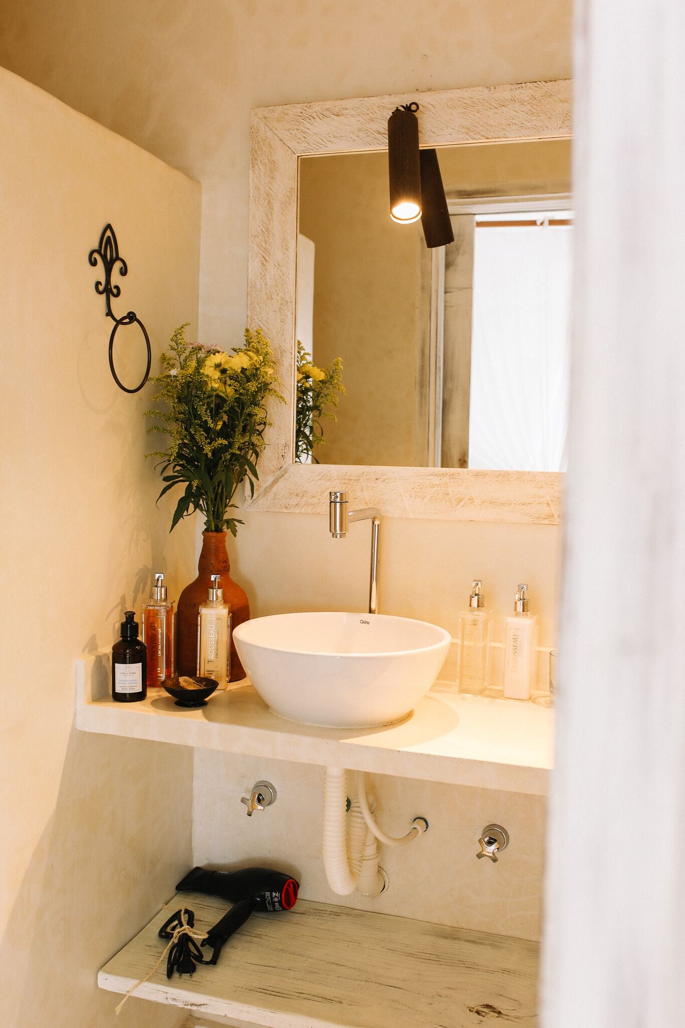 A bathroom with a white sink, a mirror with a decorative frame, a vase with flowers, and toiletries on the countertop.