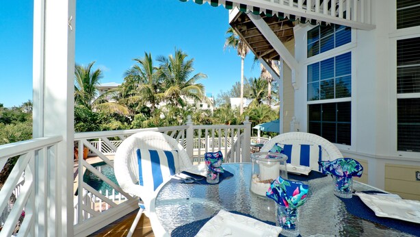 A glass-top table with striped cushions, a glass vase, and a white table runner on a balcony with white railings and palm trees in the background.