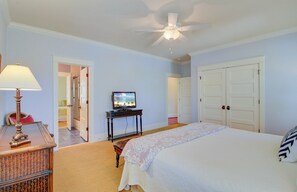 A bedroom with a bed, a television on a stand, a ceiling fan, and a door leading to another room.