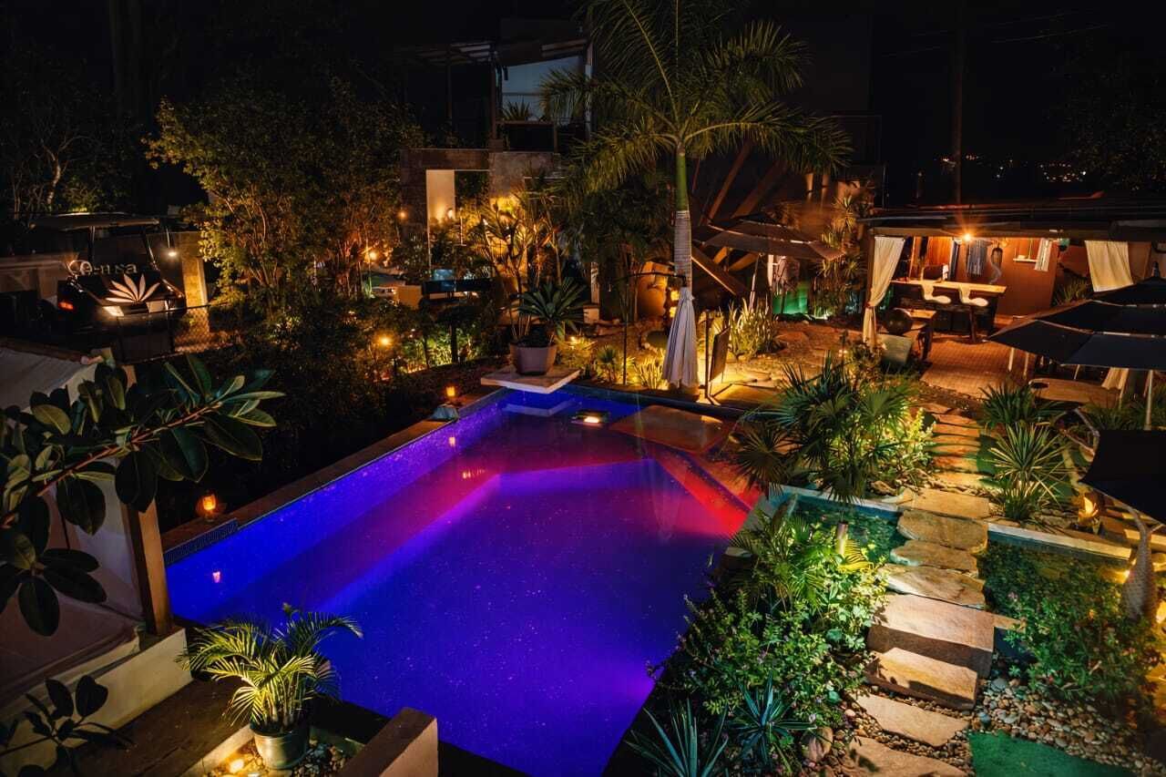 Ein Nachtbild von einem Poolbereich mit umgebender Bepflanzung und einem Gebäude mit Außenbestuhlung.