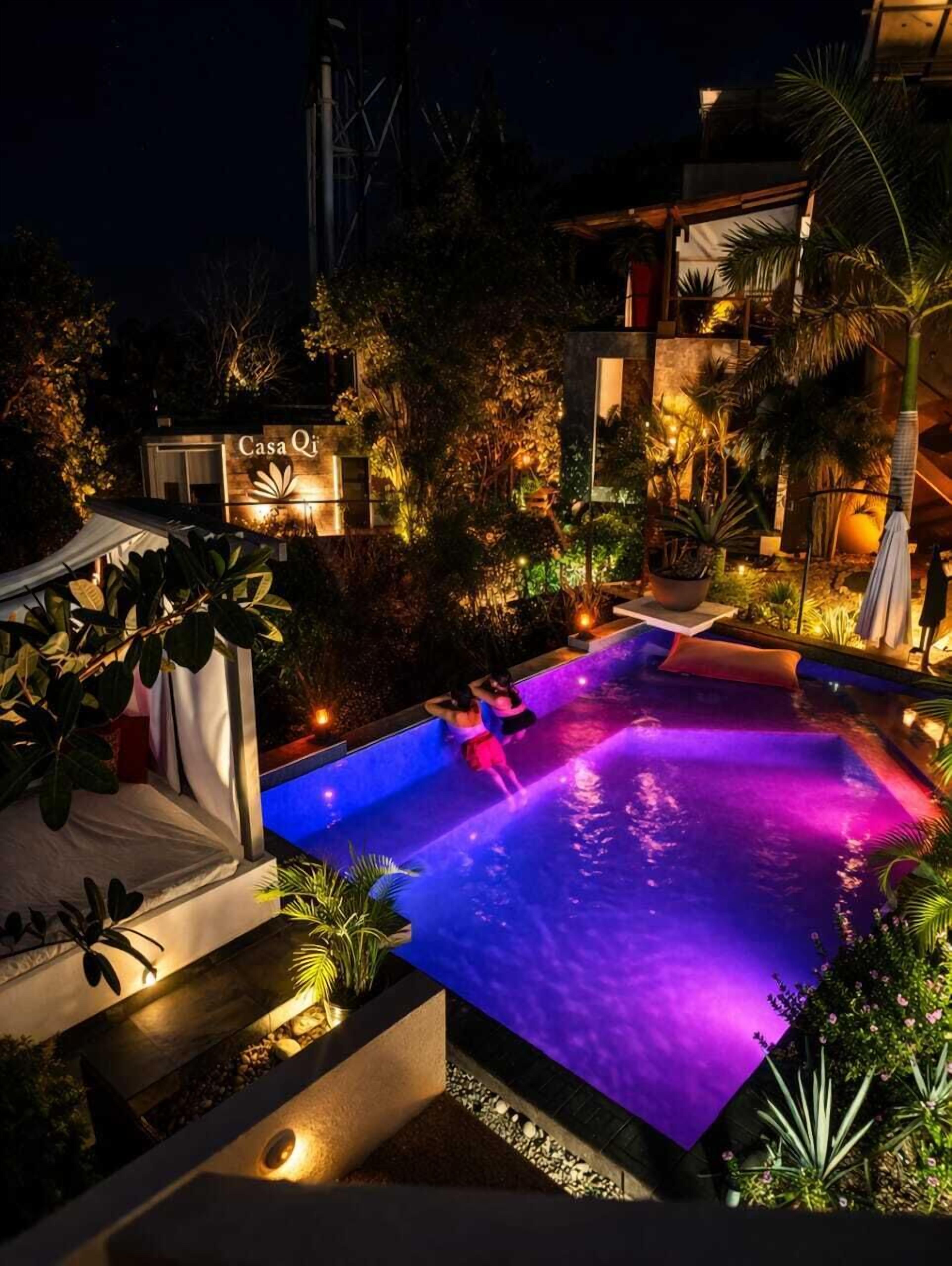 A nighttime view of a swimming pool with a sign reading 'Casa Qi' and surrounding lush greenery.