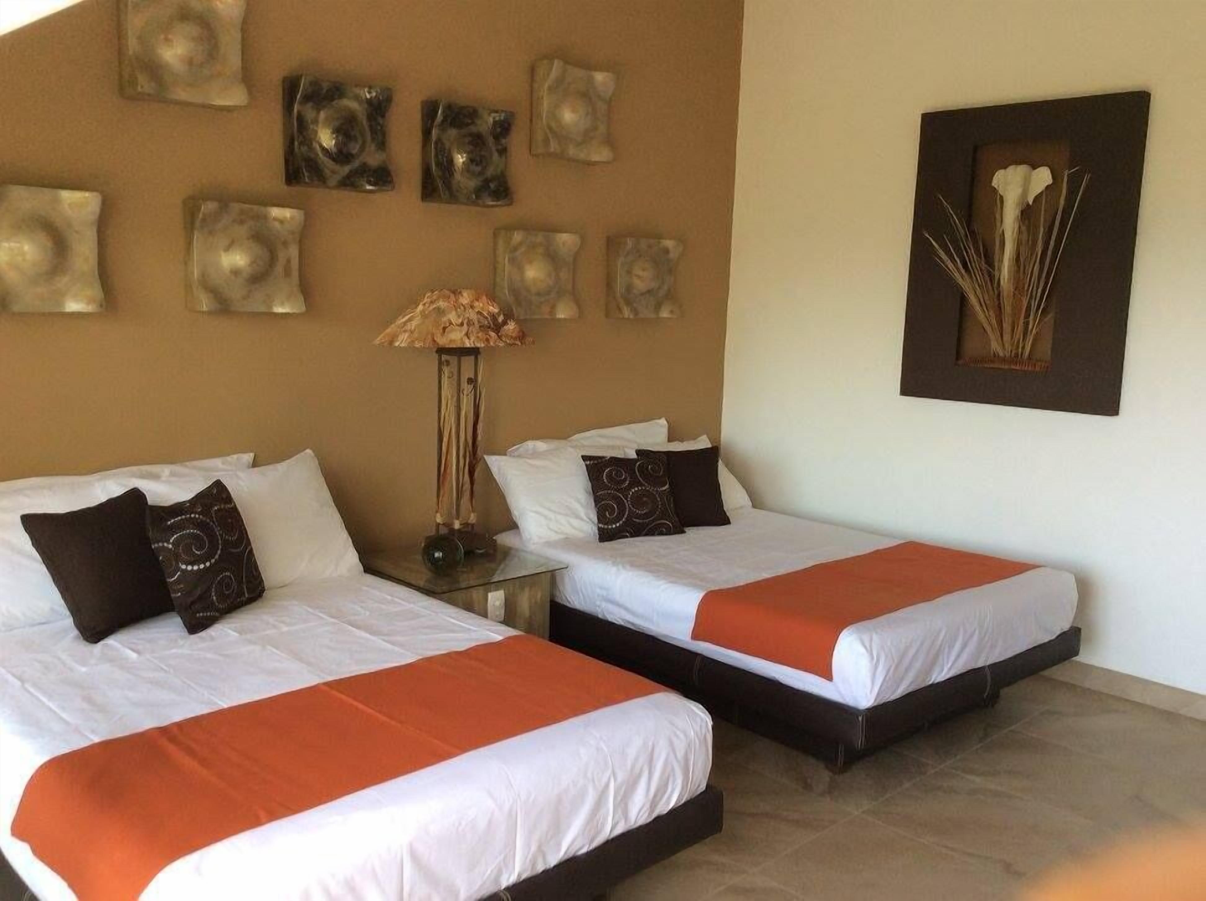 A hotel room with two beds, each with white bedding and orange accents. There are decorative wall pieces above the beds and a framed artwork on the wall.