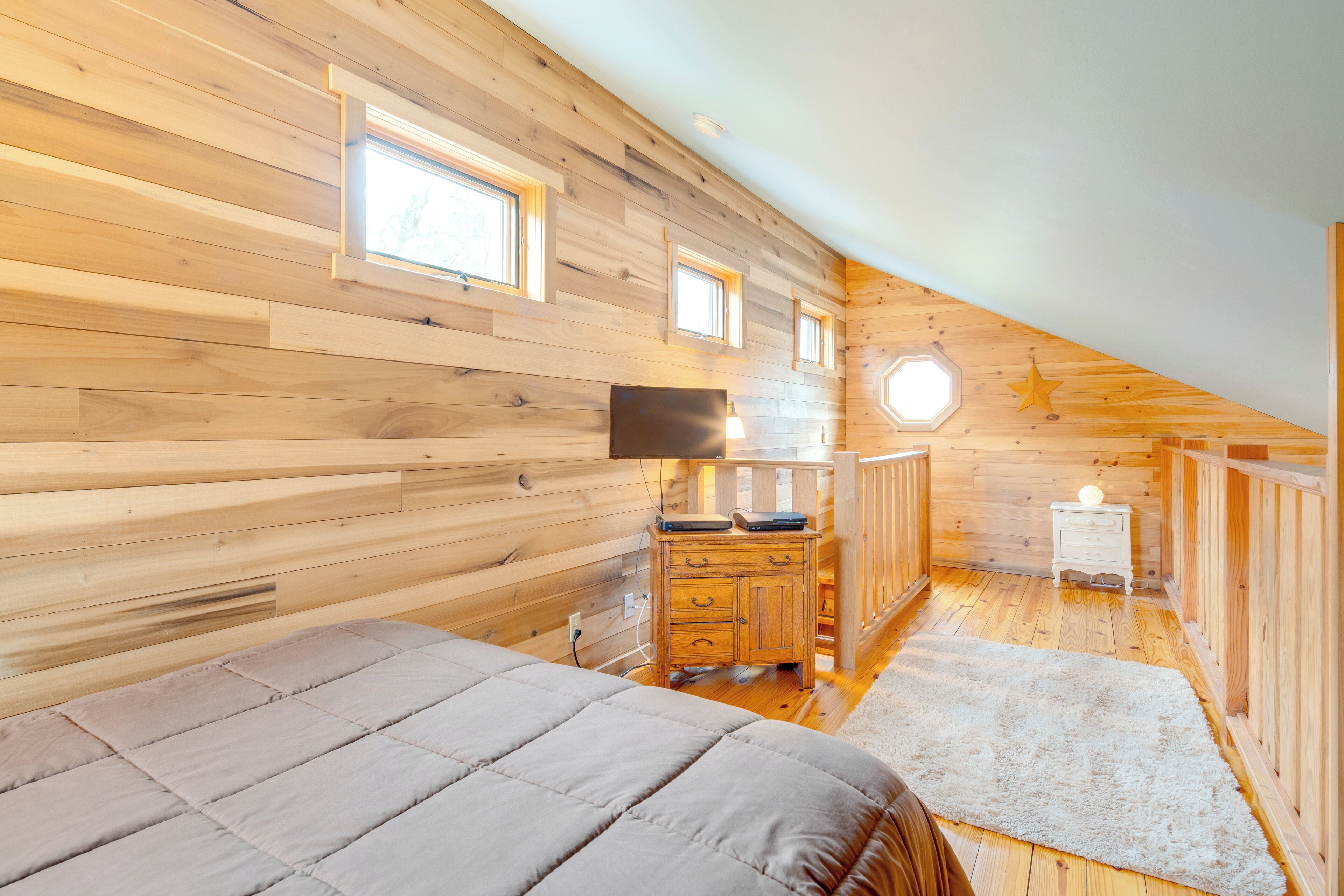 Une chambre confortable avec un plafond en lambris de bois, un lit recouvert d’une couette grise, une petite commode en bois et une table de chevet avec une lampe.