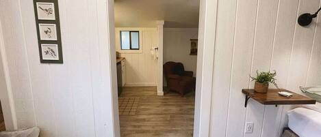 A narrow hallway with wooden flooring leading to a room with a bed and a small shelf with a plant.