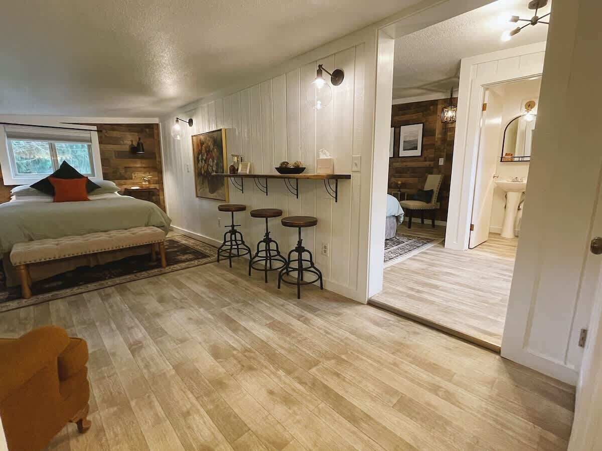 A spacious room with a large bed, a bench, and a dining area with stools. There is a kitchen visible through an open door.
