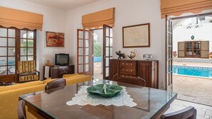 A dining area with a glass table, yellow cushioned seating, and a view of a swimming pool. - Ross Orange Apartment, Pool, Wifi (Faro)