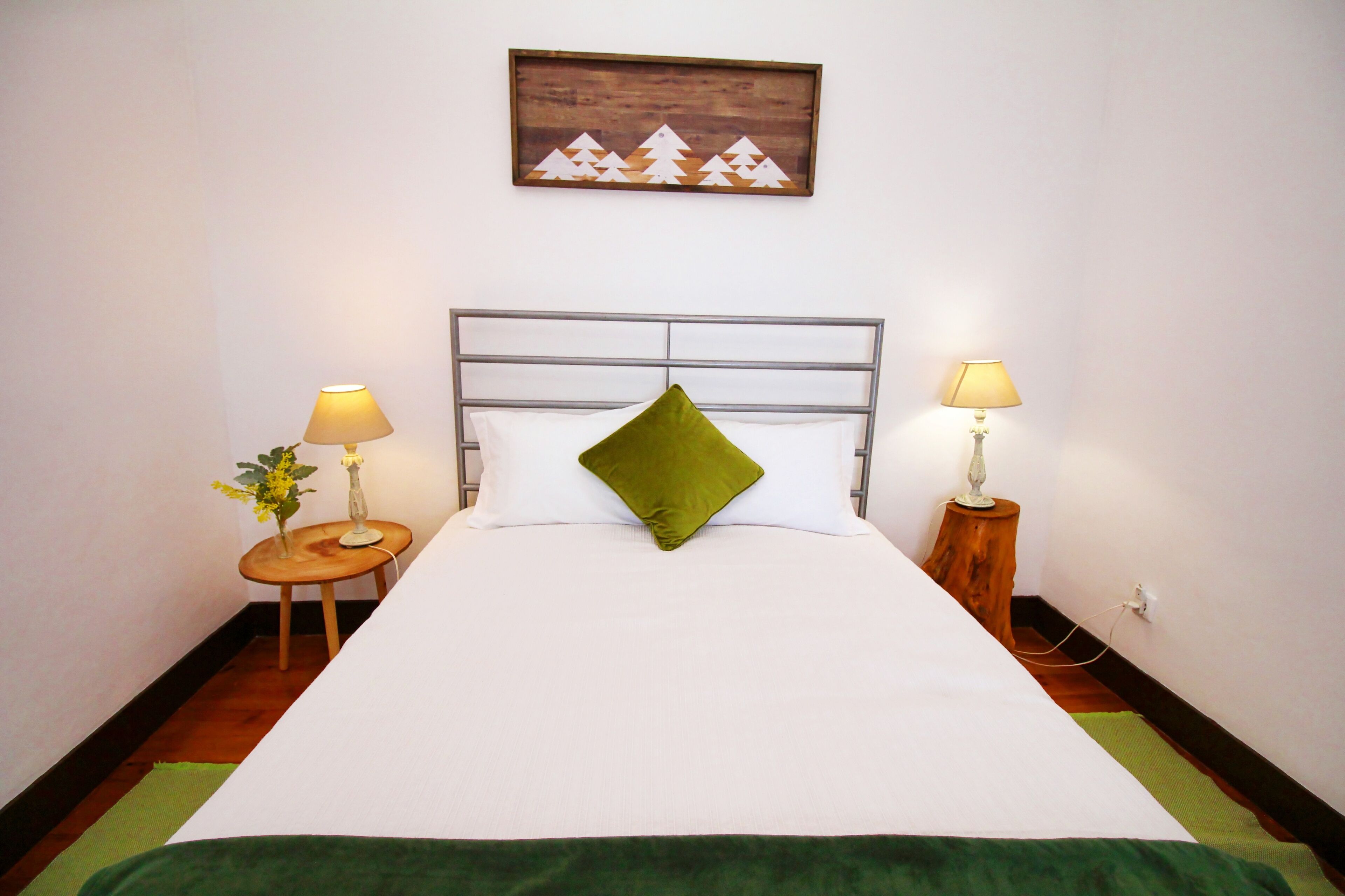 A neatly made bed with a green pillow, two bedside lamps, and a small table with a vase of flowers.