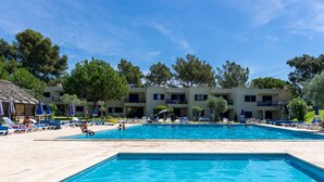 Piscina com espreguiçadeiras e guarda-sóis, cercada por um edifício com varandas e árvores.