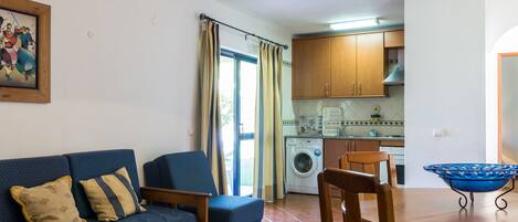 A living room with a sofa, a dining table, and a kitchen area with cabinets and a washing machine.