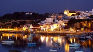 Uma vila costeira ao entardecer com barcos ancorados em um porto tranquilo, edifícios com iluminação acolhedora e uma igreja imponente em uma colina.