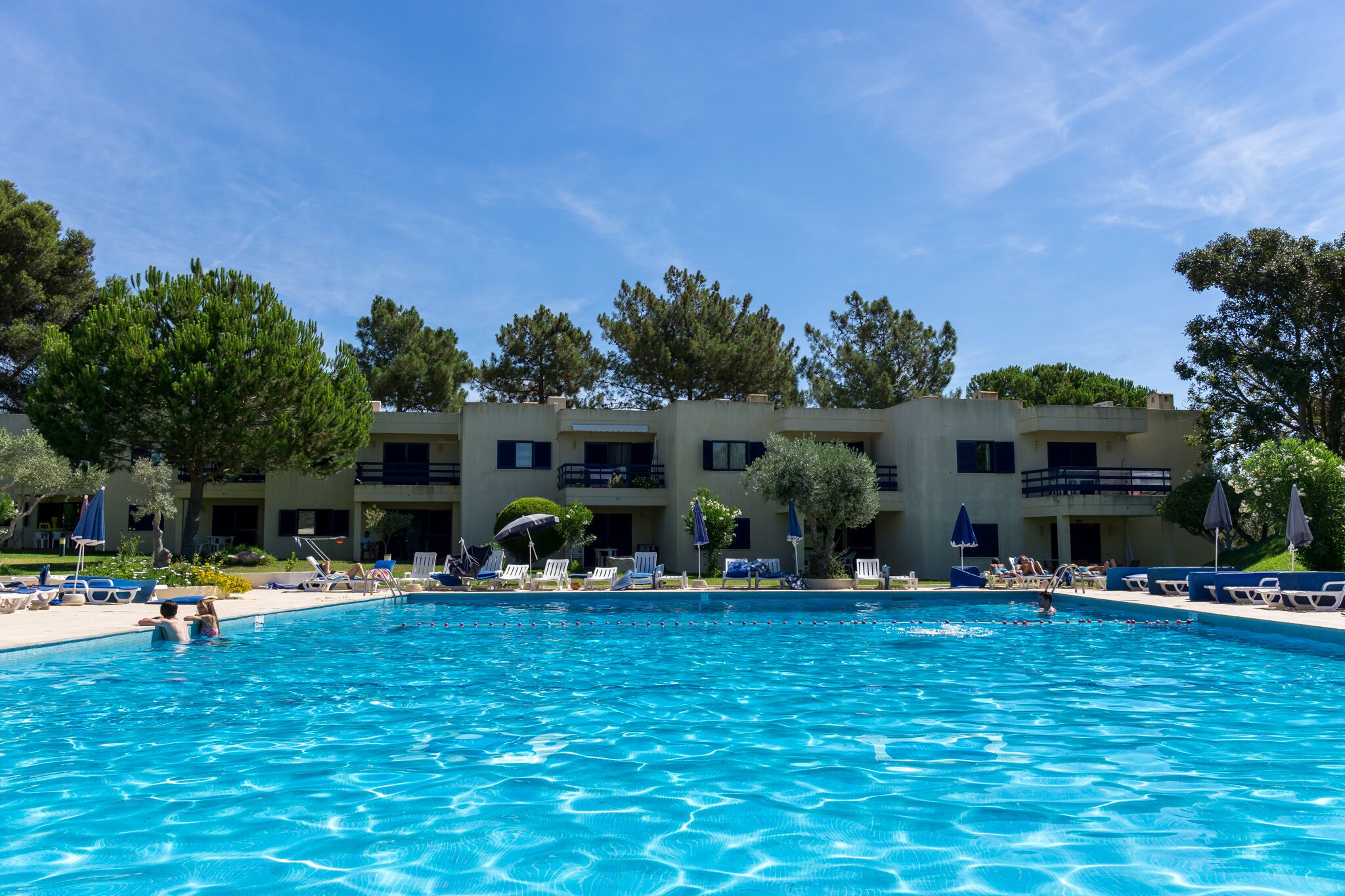 A swimming pool with lounge chairs and umbrellas, surrounded by trees and a building with balconies.