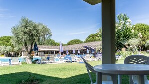 Uma área na piscina com espreguiçadeiras e guarda-sóis, um espaço coberto para sentar e um edifício com telhado de palha ao fundo.